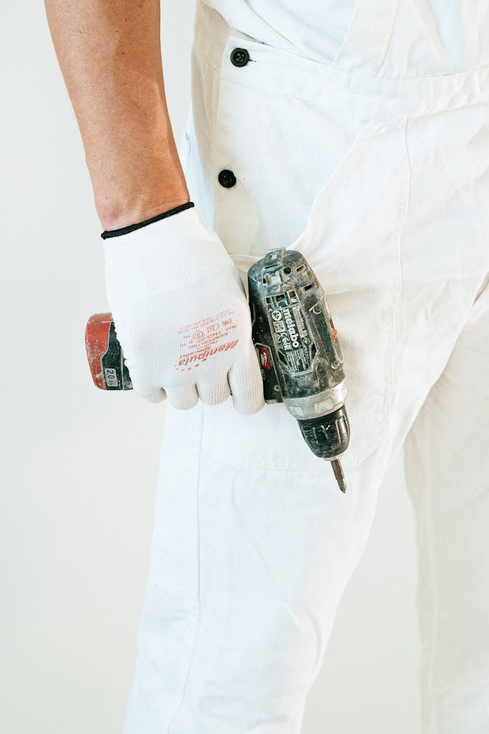 our-story Worker's hand holding power drill, wearing white overalls and gloves. Close-up view.